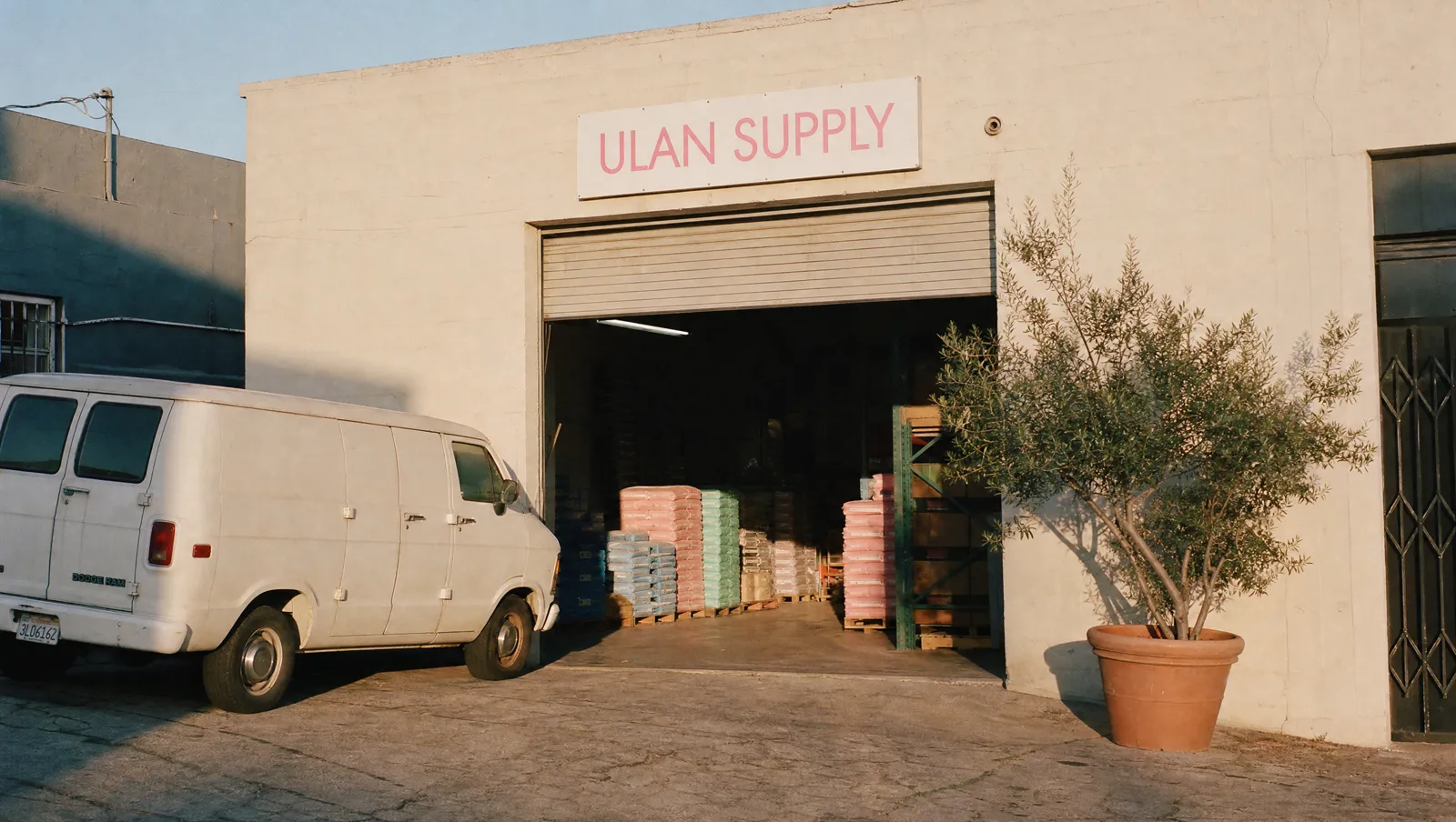 Render of the ULAN Supply factory — a small cream and kraft building with a pastel pink ULAN SUPPLY sign